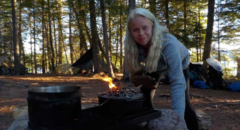 A person smiles as they prepare food over an open fire in a wooded area